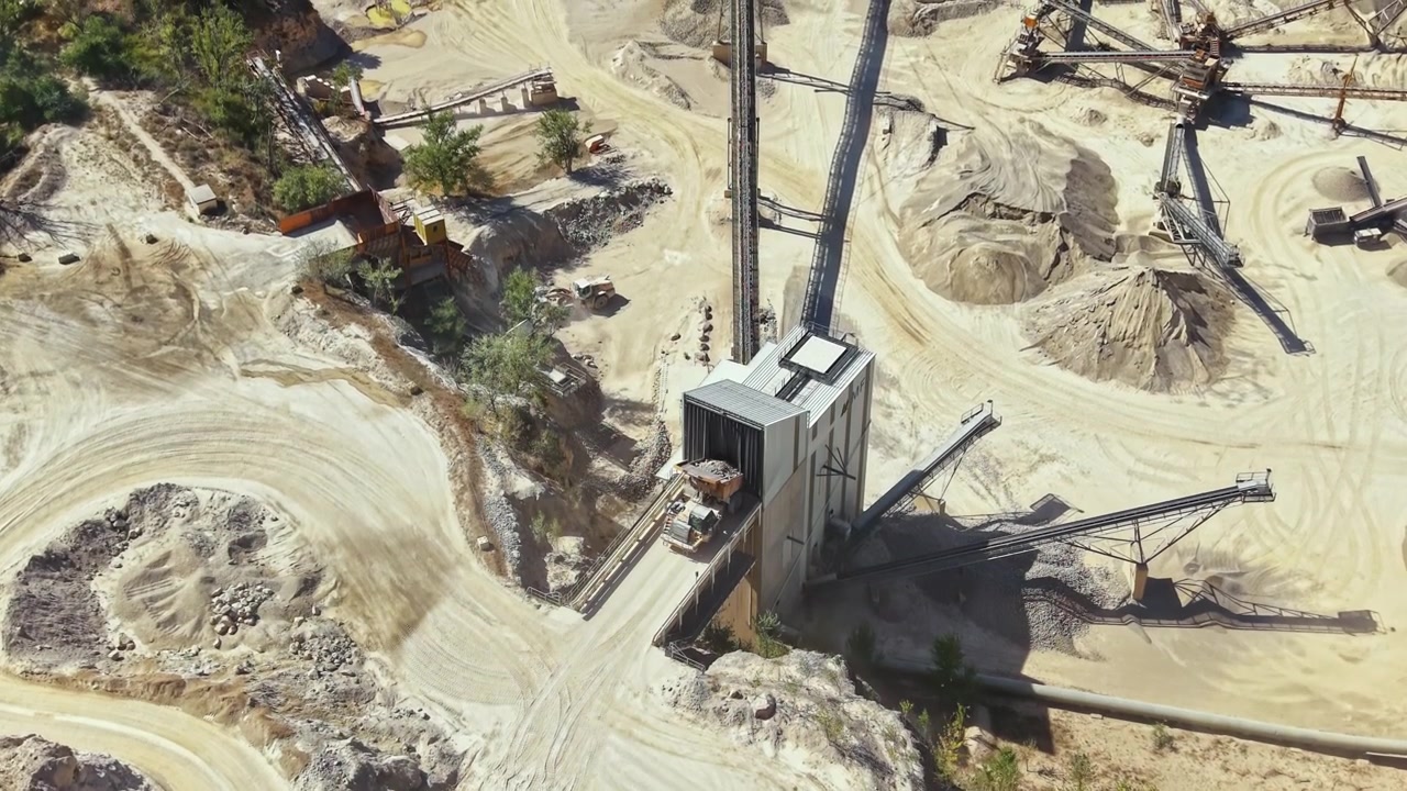 Heavy machinery operates in a sand and gravel quarry during daylight hours, with various equipment and piles of material visible from above