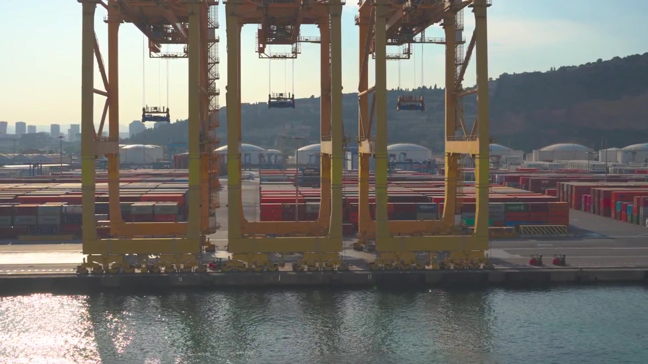 Three large yellow gantry cranes moving in sync along the quayside of a busy commercial port with rows of shipping containers in the background, showing the efficiency of modern logistics