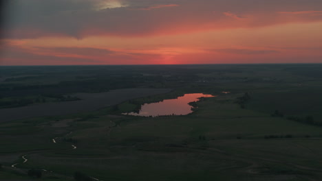 scenic landscape featuring lake reflecting vibrant red orange dusk sky above expansive flat farmland under dramatic cloud canopy with glow and winding creek creating peaceful evening vista