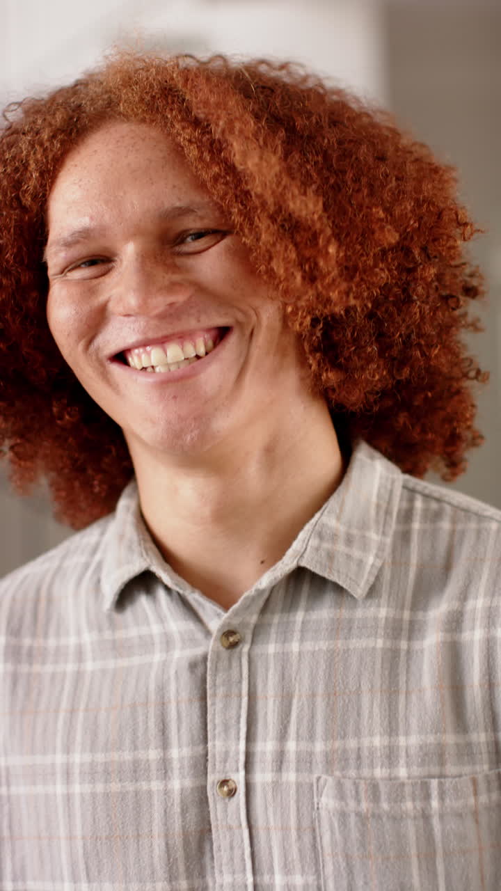 Vertical video of portrait of happy biracial man with curly hair, smiling at home, slow motion