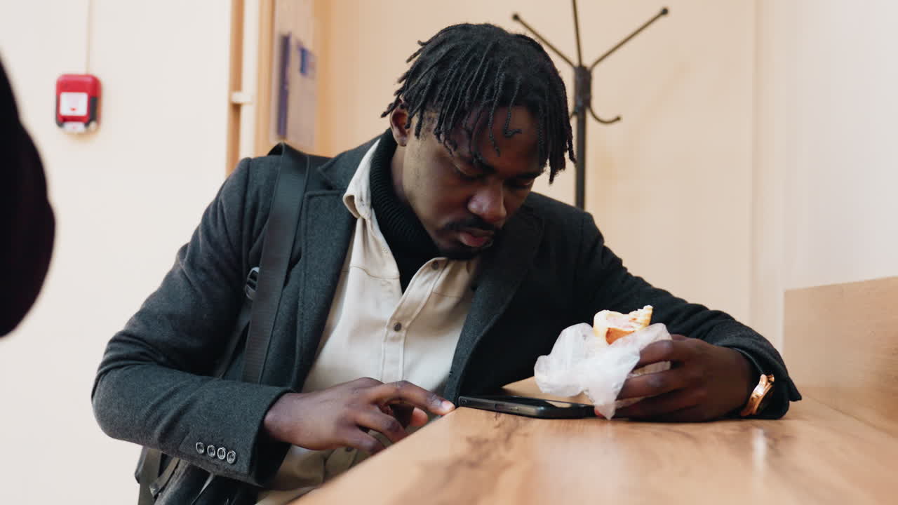 Focus on phone as dark skinned man gives mouth widening expression while eating indoors, appearing to dislodge food gently, lighting and wooden surface adding warmth to everyday realistic atmosphere
