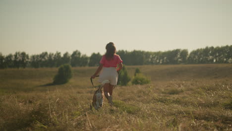 back view young girl running happily alongside her dog while holding leash in wide open farm field, with green grass and trees in background, during a bright sunny day