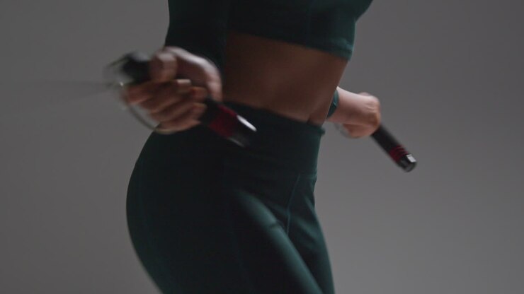 Close Up Shot Of Torso Of Female Athlete Wearing Tracksuit Training In Gym Fitness Class With Jump Rope Or Skipping Rope Against Plain Background