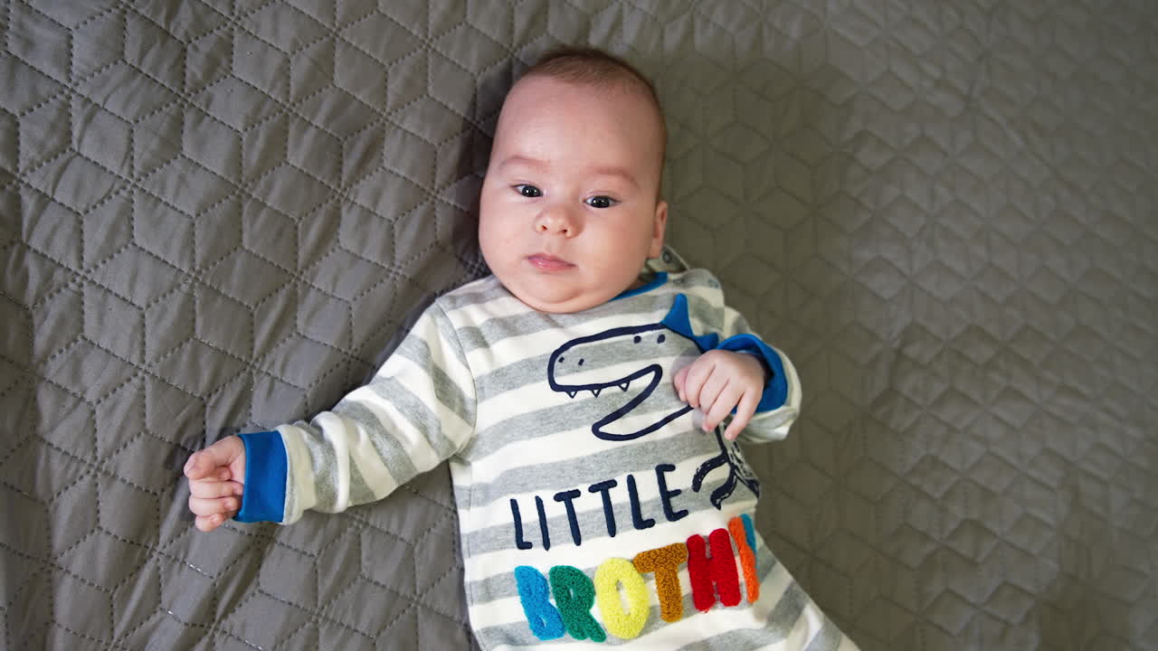 Lovely boy lying on back on a big bed. Beautiful baby wearing clothes with a little brother sign. Close up. Top view.