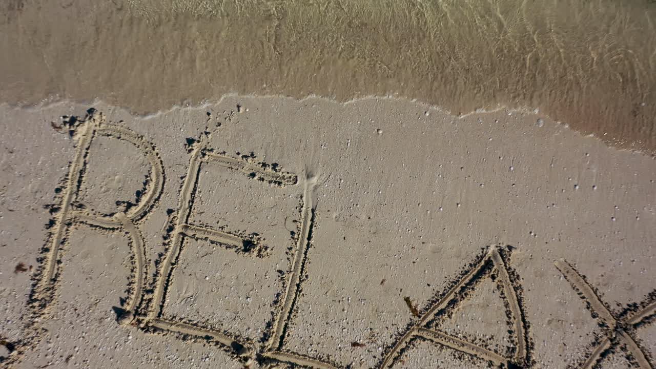 Camera rises quickly from a beach to reveal RELAX written in the sand