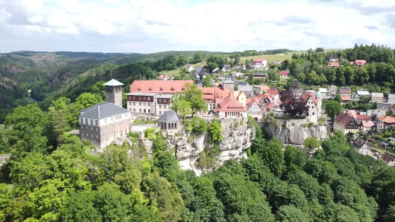 Drone moving closer to a castle on a hill with trees and village in the middle of Saxony on a bright but cloudy day