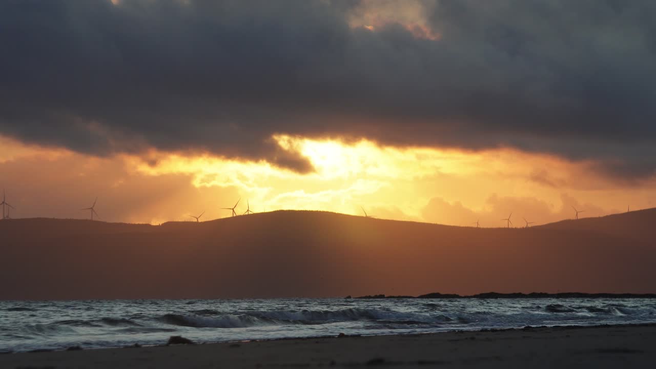 A scenic sunset over a Scottish bay with waves crashing onto shore, silhouetted mountains in the distance, and wind turbines turning on the ridgeline. A calm, atmospheric coastal scene.