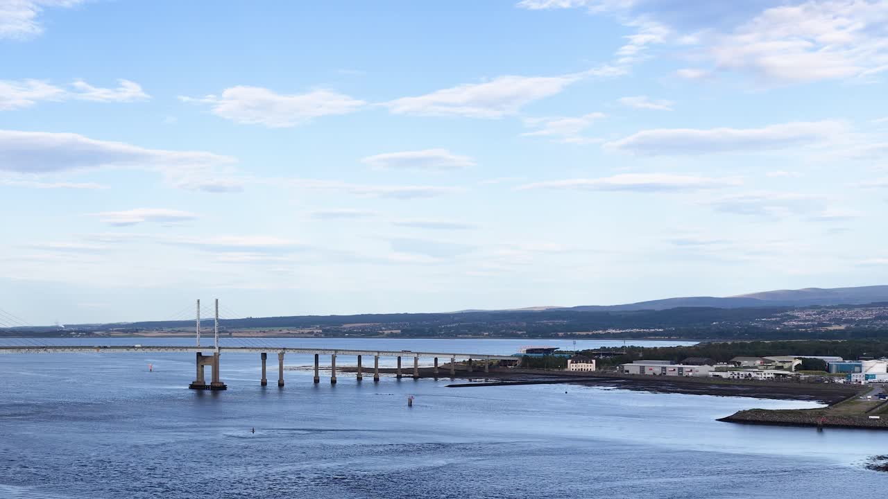 Wide aerial pan of modern bridge spanning river, under bright daylight, with distant cityscape visible