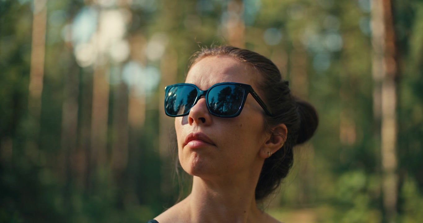 Close-up portret van een vrouw met zonnebril in een zonovergoten dennenbos. Warm natuurlijk licht en een zacht vervaagde bosachtergrond creëren een ontspannen buitenleven scène