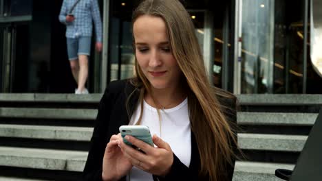 attractive businesswoman looking at smartphone screen near city building