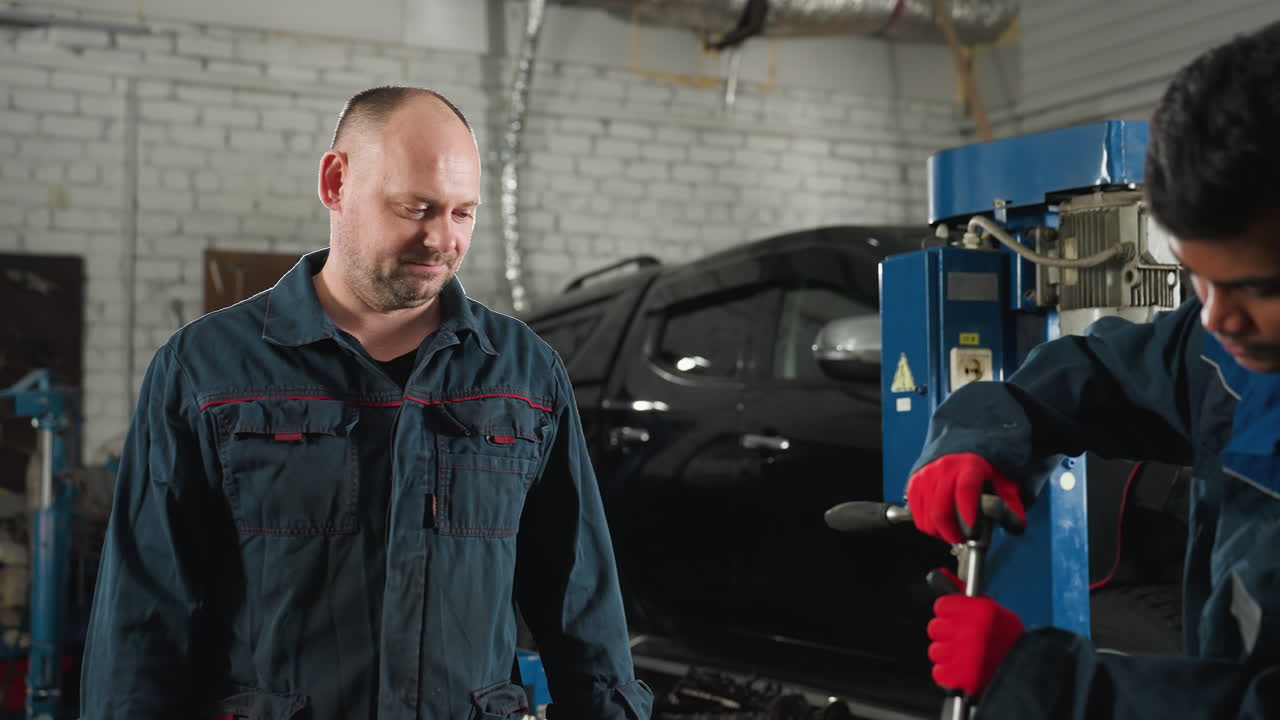 ingenieros de automóviles en un taller de automóviles, uno con guantes rojos trabajando en un motor de automóvil usando una herramienta mientras un colega observa, el fondo presenta coches estacionados, equipos de taller