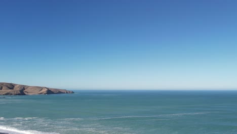 High-altitude-slow-reverse-above-beautiful-turquoise-ocean-with-sloping-land,-incoming-waves-and-stony-beach--Birdlings-Flat-Beach,-New-Zealand