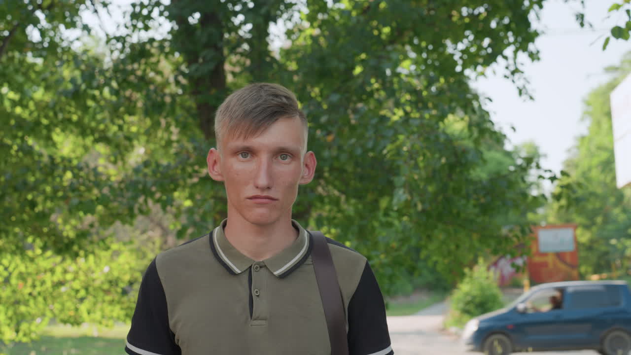 Frontal Outdoor Portrait Under Dappled Foliage, Young Man Faces Camera With Serious Neutral Expression, Backpack Strap And Quiet Pathway Visible, Warm Natural Light And Soft Shadows