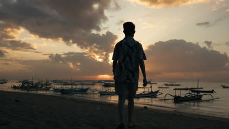 Silhouette-Eines-Mannes,-Der-Zum-Sonnenuntergang-Am-Strand-Mit-Bootshafen-Geht