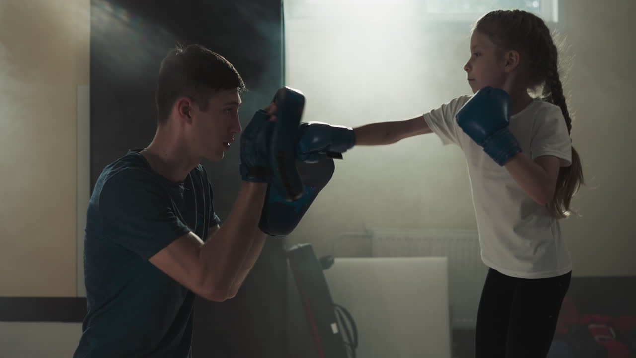 아이는 체육관에서 트레이너와 스파링하는 동안 패드를 때립니다. 권투 강사는 스포츠 학교에서 작은 소녀와 전투를 수행합니다. 느린 움직임. 신체 활동