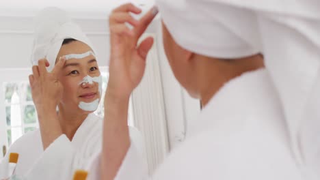 video of happy asian woman in robe with moisturizing mask looking at mirror