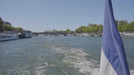 Vista-De-La-Ciudad-Y-Los-Puentes-Desde-Un-Barco-Turístico-Por-El-Río-Sena-En-París,-Francia,-Con-Bandera-En-Primer-Plano