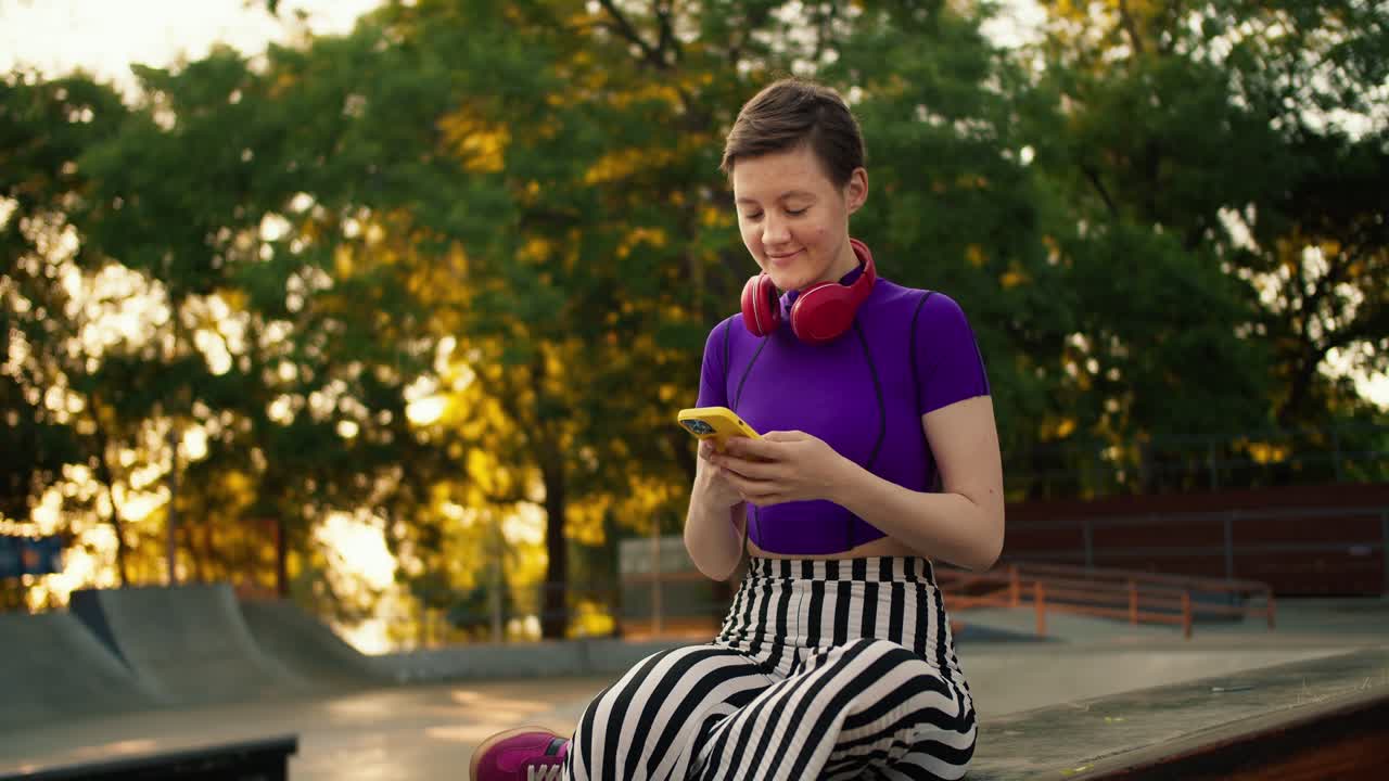 una chica joven con un corte de pelo corto en una camiseta púrpura se sienta en las redes sociales y se toma una selfie usando un teléfono amarillo en un parque en verano. adolescente con auriculares rojos en el parque