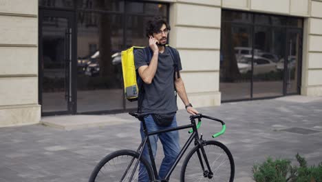 delivery man with yellow backpack walk on the street with a bike, using earphones answer to a call