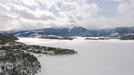 aerial moving backward to reveal icy, frozen lake surrounded by snow and green pine trees with large snowy ski town mountains in the background near silverthorne and frisco colorado