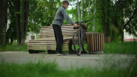 boy in ash top rides bicycle through park, stops by wooden bench, parks bike between bench and nearby wooden structure, sits down and joins hands together, surrounded by green grass and trees