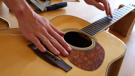 close up hands of a luthier craftsman measuring and leveling an acoustic guitar neck fretboard on a wood workshop bench with lutherie tools