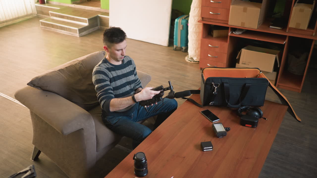 Man adjusts microphone on camera, preparing filming setup in home studio. Organized workspace with camera gear, tools, and lighting equipment on wooden table. Focused on creative process
