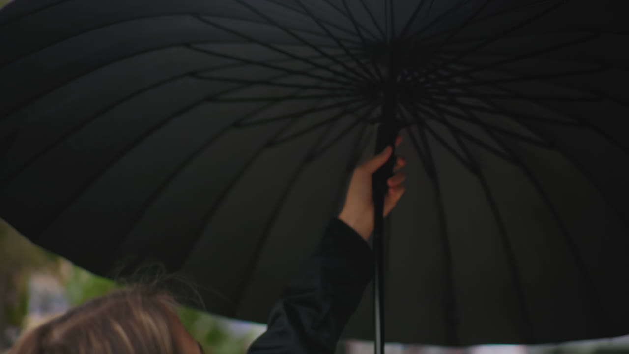 Lady in black coat smiling while opening an umbrella on city street with blurred background of trees buildings and lamppost during cloudy weather showing lifestyle mood moment in urban environment