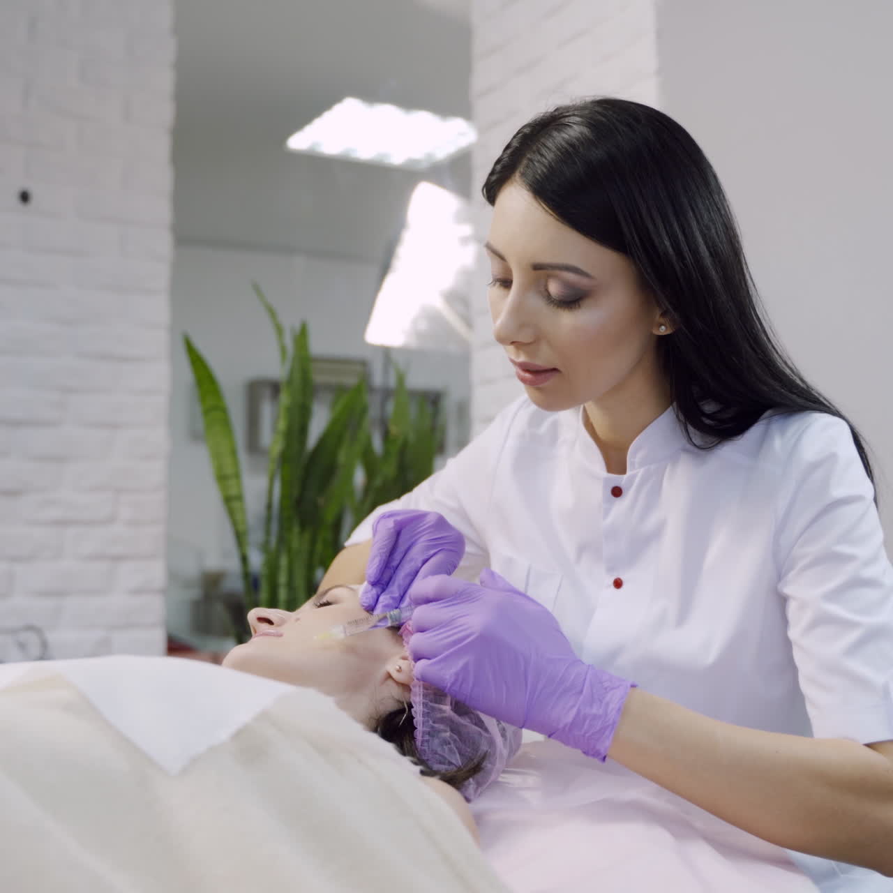 A beautician with long hair checks an injection and starts the biorevitalization procedure on the woman's face. Blurred background.