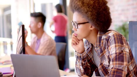 mujer biracial pensativa usando la computadora en el escritorio en una oficina casual, cámara lenta