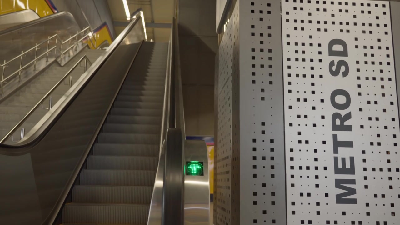 Et panorameskud af et moderne metrostationinteriør med en opadgående rulletrappe, betontrapper og et Metro SD-søjleskilt.