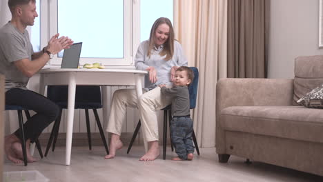 Rear-View-Of-A-Baby-Taking-His-First-Steps-Towards-The-Table-Where-His-Parents-Are-Sitting