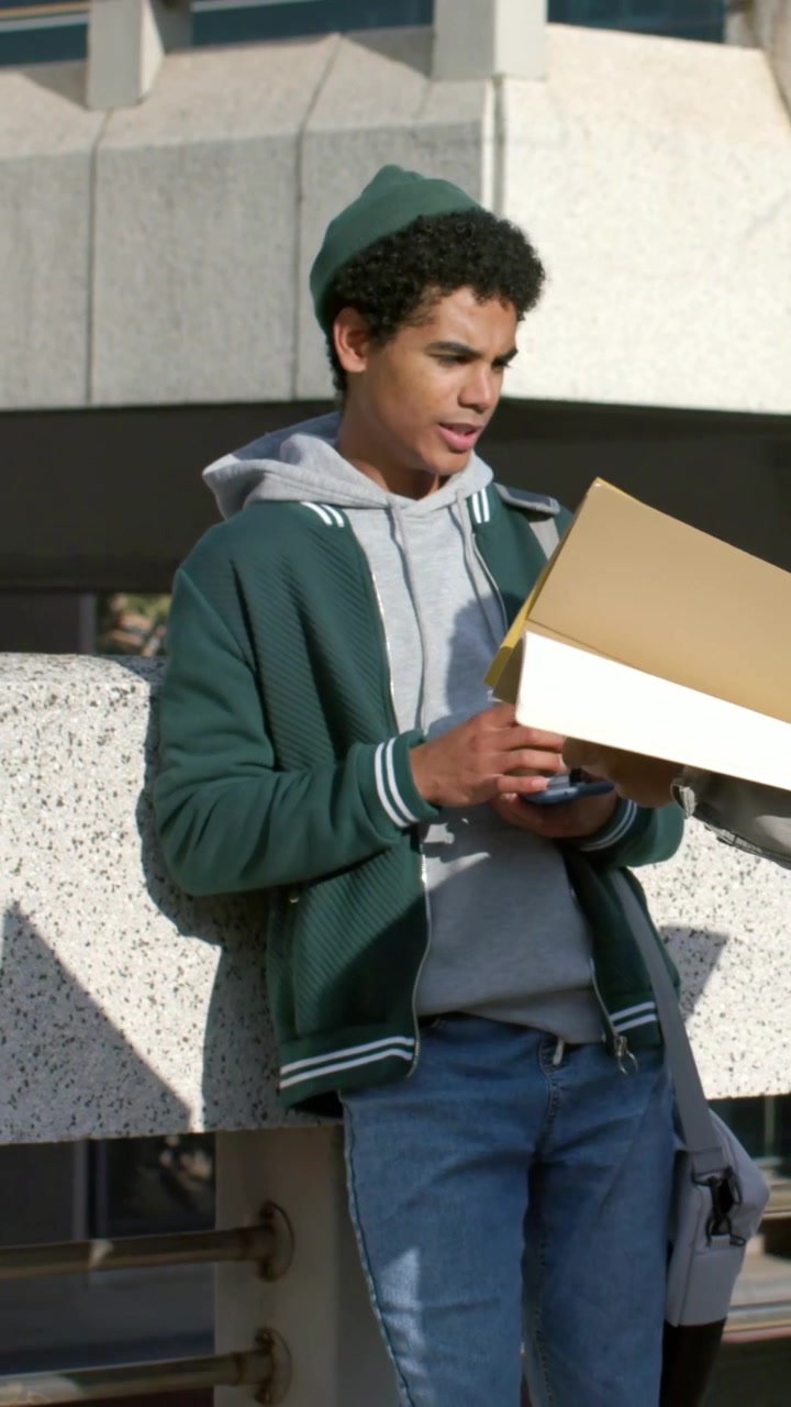 Video verticale: Uomo appoggiato con cappello verde che tiene una tazza, usa il telefono nel campus, prende una cartella da un collega