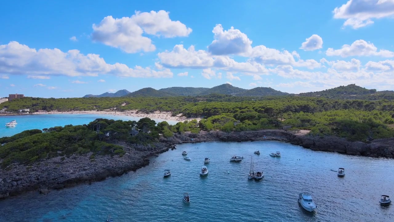 Viste mozzafiato di Cala Agulla mostrano una costa serena con barche che punteggiano le acque limpide e blu. Una vegetazione lussureggiante e montagne distanti creano un paesaggio pittoresco perfetto per il relax.