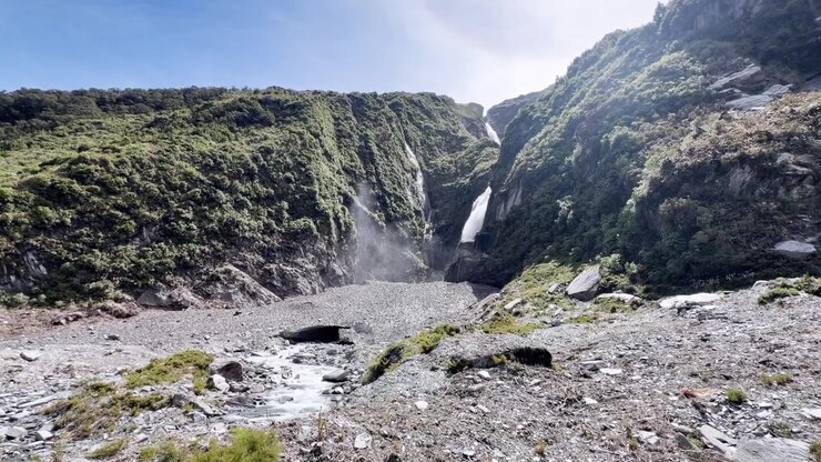 Powerful alpine waterfall plunging down steep rocky cliffs on a bright sunny day in New Zealand. Crystal-clear glacier-fed water flows through a rugged mountain landscape beneath clear blue skies.