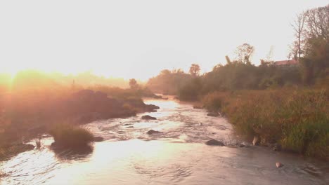 drone video with moving forward along a river with morning sun rays glittering off the water on a foggy morning