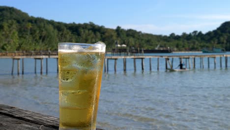 A-closeup-of-a-beer,-on-the-beach,-slightly-angled-with-a-pier-in-the-background