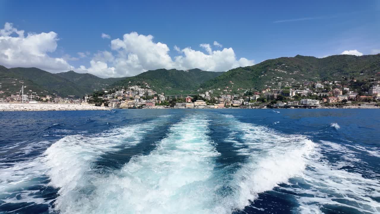 Vista grandangolare da una barca in movimento che cattura vivaci scie sull'acqua blu profondo, con un'incantevole città costiera e rigogliose colline verdi in pieno giorno, ricco contrasto cromatico.