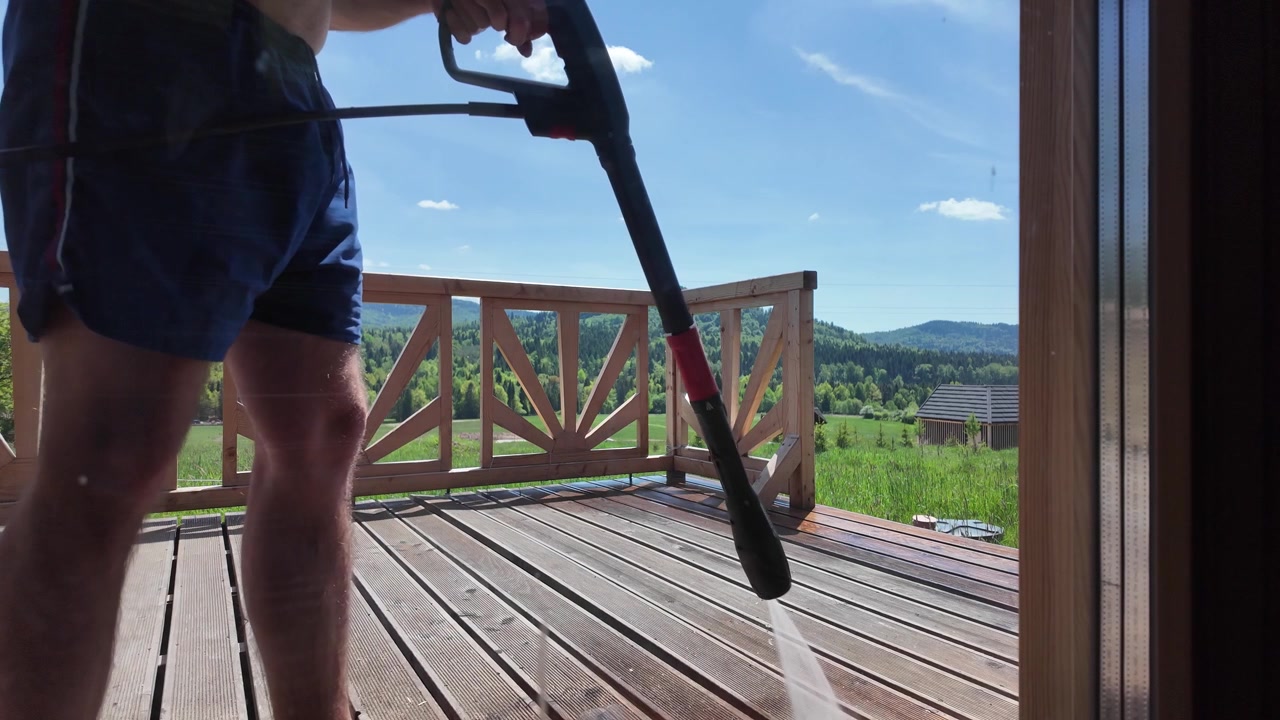 Shirtless worker in blue shorts cleans a wooden terrace with a pressure washer, with green grass, forest and mountain peaks visible in the background.