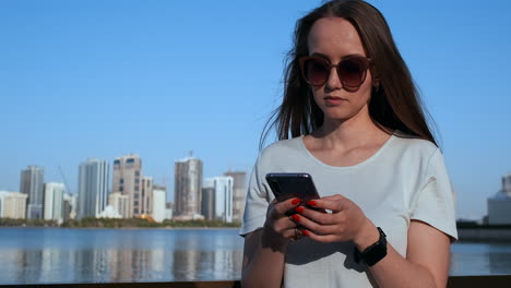 Hermosa-Chica-Con-El-Pelo-Largo-Con-Gafas-De-Sol-Usando-Una-Aplicación-Para-Teléfonos-Inteligentes-En-El-Muelle-Del-Río-Sunset-Cerca.