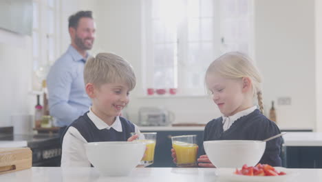 two children wearing school uniform in kitchen eating breakfast as father gets ready for work