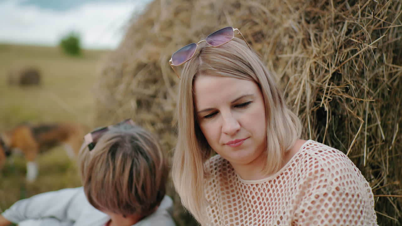 задумчивая женщина с солнцезащитными очками на голове смотрит вниз в спокойной сельской обстановке, другая женщина сидит рядом, с размытой собакой на заднем плане, рядом с большим пакетом сена на обширной открытой сельскохозяйственной земле