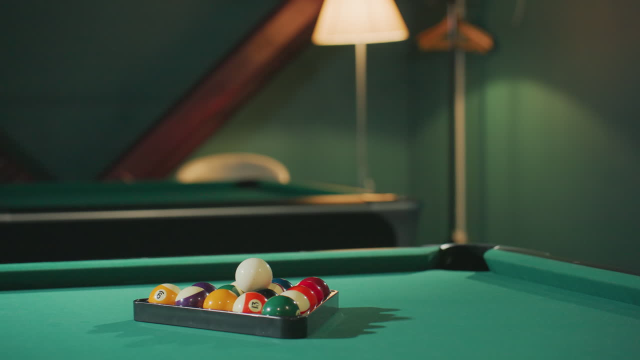 Vibrant billiard balls arranged in triangle rack on green pool table under warm lighting in cozy game room. Classic cue sport setup with focus on white cue ball and numbered balls in dim ambiance