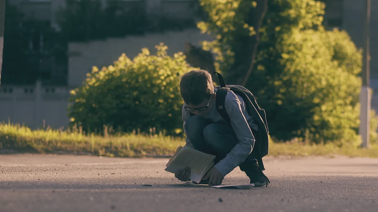 un ragazzo con i capelli biondi raccoglie fogli di carta e libri sulla strada