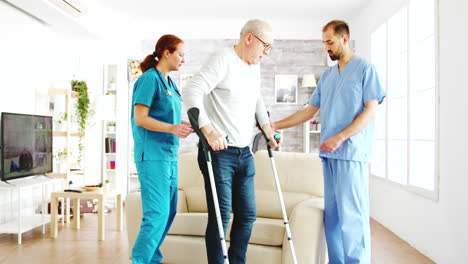 two nurses helping an old disabled man with crutches to walk in his room