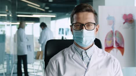 close-up view of handsome young male doctor in glasses sitting in hospital office at work, looking at camera and smiling