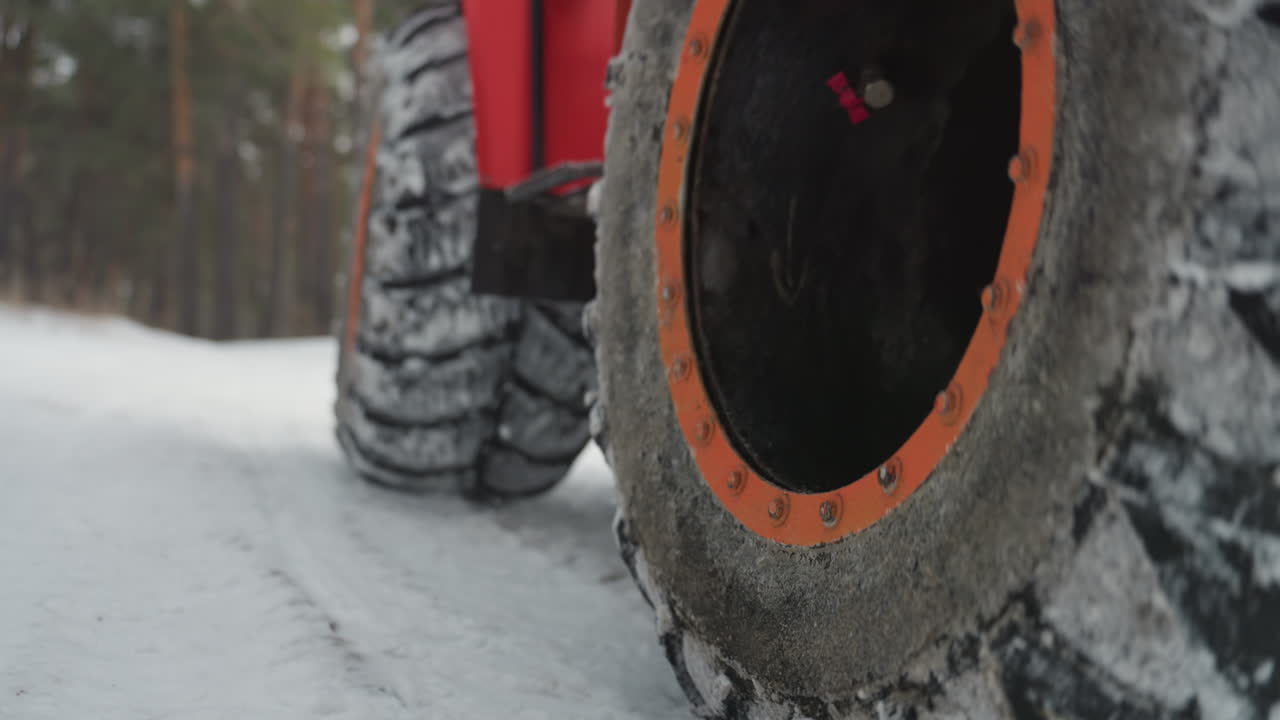 Red atv tires on icy road, lowangle closeup showing heavy tread packed with snow, orange rim and rugged suspension, pine forest backdrop, cold gritty atmosphere conveying extreme offroad grip