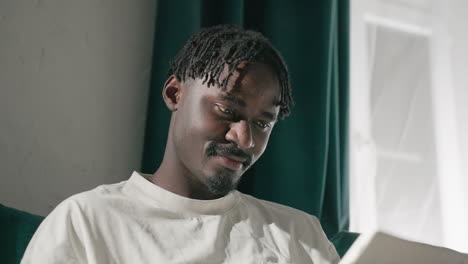 black youth smiling while using tablet on teal sofa, looking content and relaxed. soft natural light from the window highlights his joyful expression, creating a warm, peaceful moment at home