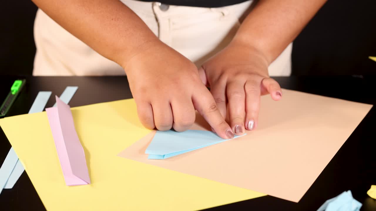 Person folds blue origami paper on yellow sheet under bright lighting, camera overhead, steady view