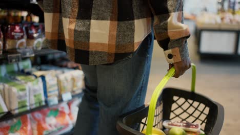 un hombre con una camisa a cuadros con color de piel negra y vaqueros azules camina a lo largo de los mostradores en un supermercado moderno y pone los productos seleccionados que necesita en su cesta de comestibles